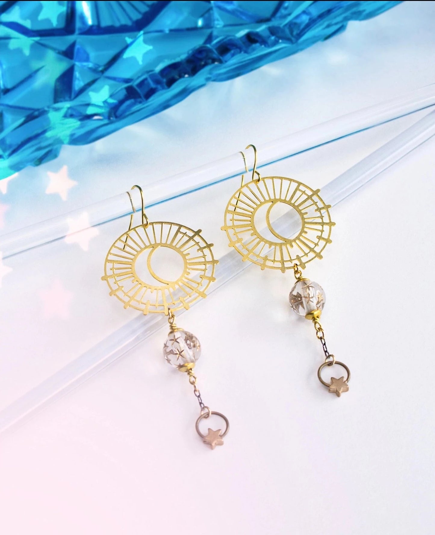 This photo shows the earrings hanging from a clear jewelry display with some blue in the top of the background. Earrings are gold in color with a clear bead dangling, and another star dangling from the bead