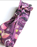 This photo shows the hair scarf tied in a know at the top of it. This photo is also at an angle. This scarf has different mermaids with dark colored tails, pink whales, and various sea plants on a dark purple background