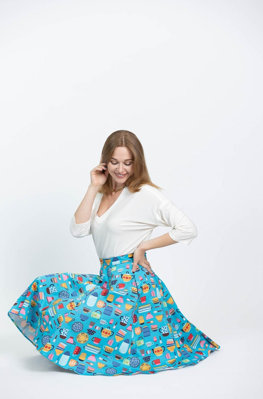 This photo shows a white female presenting model wearing a white blouse with the Kitchenware skirt. Skirt has a array of vintage kitchen items such as mixing bowls, tea kettles, mugs, etc on a light blue background.  Model is shown crouching close to the ground and looking down at the skirt.