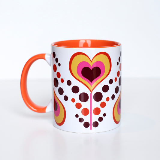 This mug has an orange, dark maroon, yellow, and pink coloring. Hearts have alternating colors inside of each other and are on a pink stem. Each heart flower alternates being upright and upside down with pink, maroon, and yellow dots as leaves flowing between each heart flower. Mug handle, and inside coloring is orange