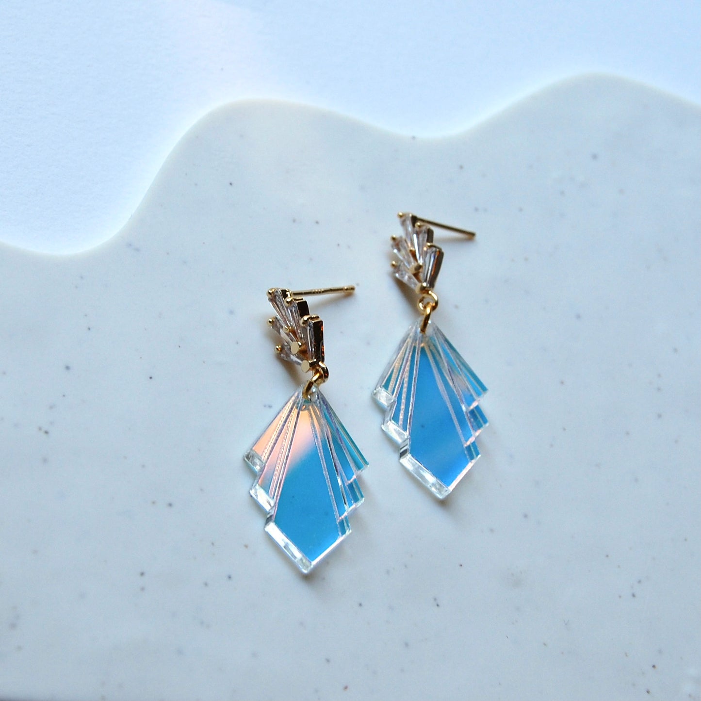 Photo shows the product laying on a layered surface. Earrings have 5 rectangular cubic zirconia stones in a fan at the post, and has a fan in an acrylic iridescent style for the bottom of the earrings