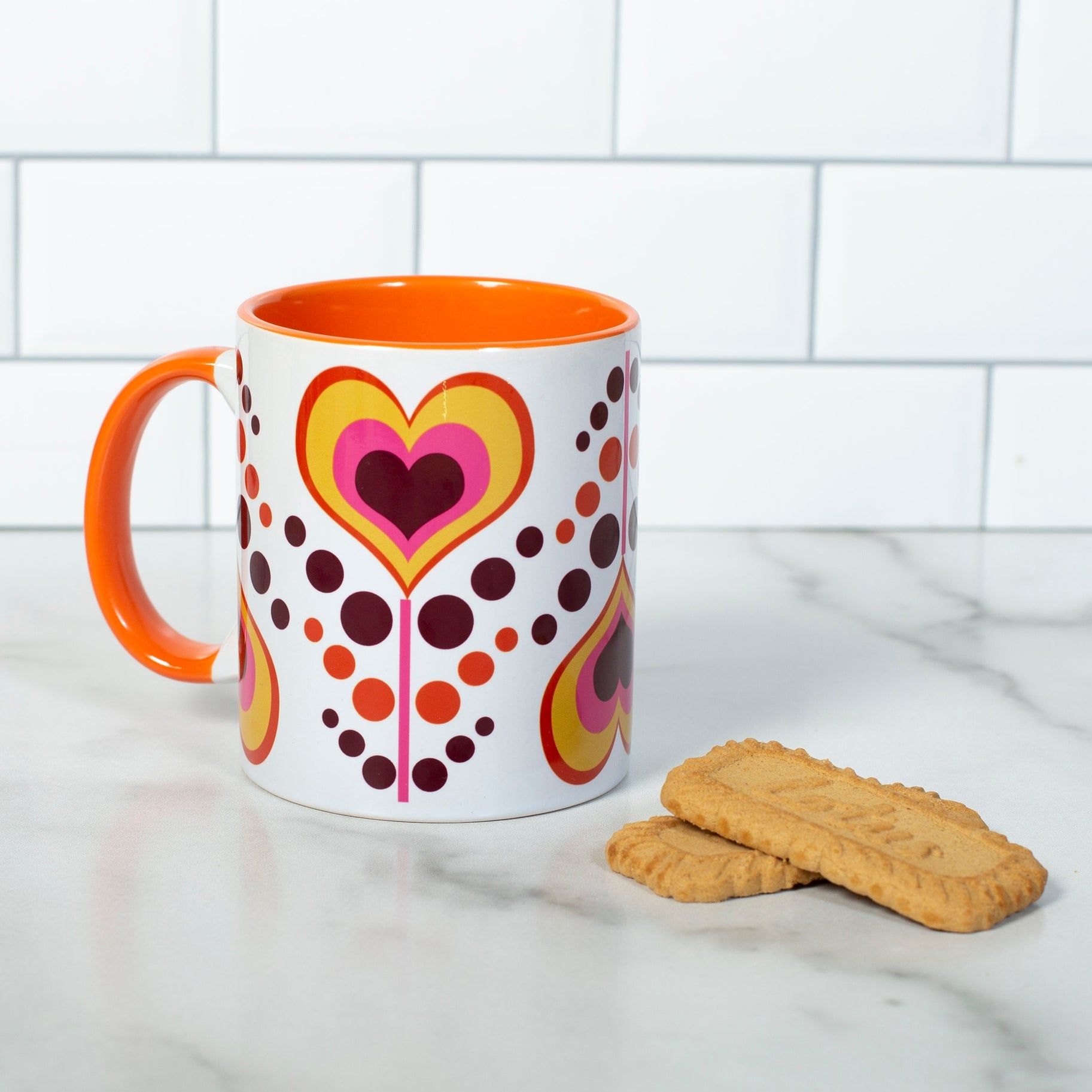 This photo shows the mug next to 2 plain cookies. This mug has an orange, dark maroon, yellow, and pink coloring. Hearts have alternating colors inside of each other and are on a pink stem. Each heart flower alternates being upright and upside down with pink, maroon, and yellow dots as leaves flowing between each heart flower. Mug handle, and inside coloring is orange