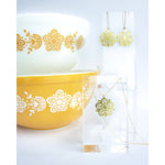 This photo showcase the Pyrex Flower earring and necklace next to a large yellow pyrex bowl and a small white pyrex bowl, each with the same flower pattern.
