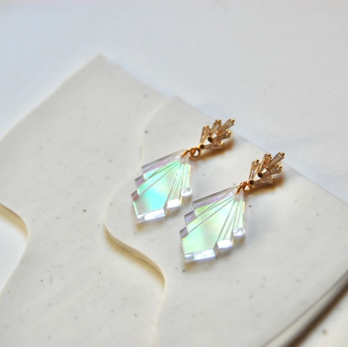 Photo shows the product laying on a layered surface. Earrings have 5 rectangular cubic zirconia stones in a fan at the post, and has a fan in an acrylic iridescent style for the bottom of the earrings