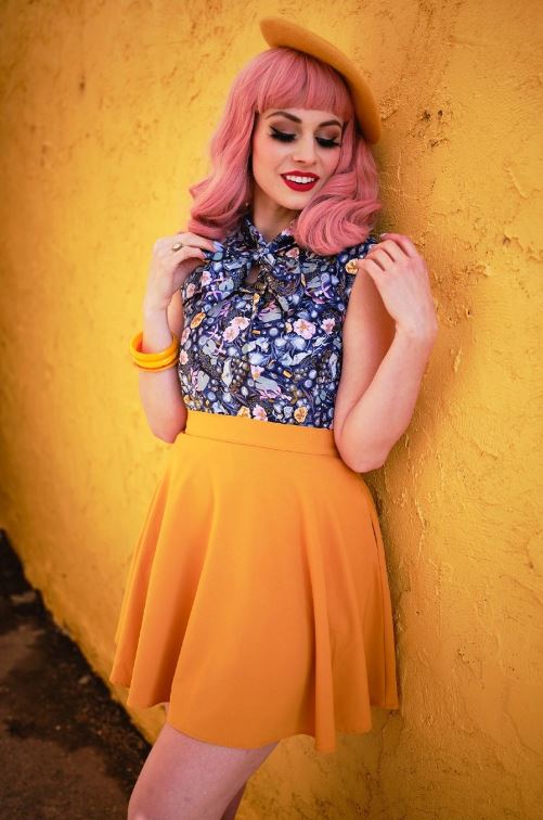 This photo shows a female presenting model wearing the skirt against a yellow background. Skirt is above the knee. Model has pink hair, and is wearing a blue top