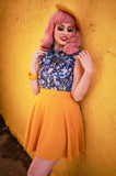 This photo shows a female presenting model wearing the skirt against a yellow background. Skirt is above the knee. Model has pink hair, and is wearing a blue top