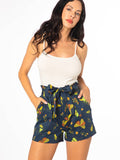 This photo showcases a white female presenting model wearing a white tank top and the Summer Night shorts. Shorts are a dark blue with yellow and gold colored insects including moths, and beetles. Also includes flowers.