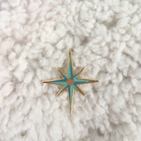 Photo shows an upclose look at the starburst pin. Pin is dark blue with light blue accents, and a mirror like circle in the center of the pin. Pin is shown against a white carpet like backgound