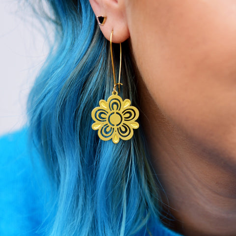 A close up of the dangle earring. Earring is gold in color, and is a metal cutout of a Pyrex flower. Earring is show on a white female presenting person with blue hair