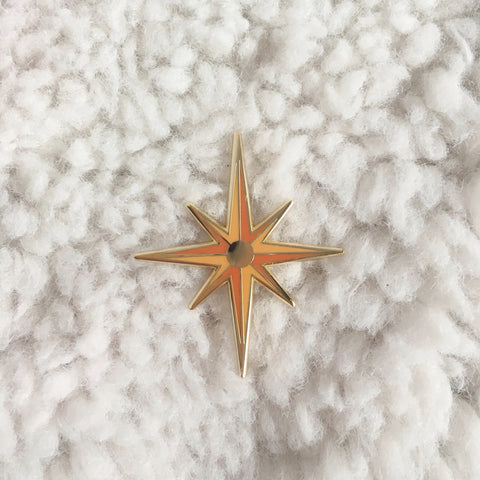 Photo shows an upclose look at the starburst pin. Pin is dark orange with light orange accents, and a mirror like circle in the center of the pin. Pin is shown against a white carpet like background