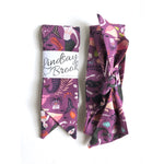 This photo showcases 2 hair scarves next to each other. One in the packaging, and one with a bow. This scarf has different mermaids with dark colored tails, pink whales, and various sea plants on a dark purple background