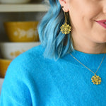 This photo shows the female presenting person wearing the Pyrex Flower earring and necklace. Person is white with blue hair, and wearing a blue sweater