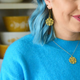 This photo shows the female presenting person wearing the Pyrex Flower earring and necklace. Person is white with blue hair, and wearing a blue sweater