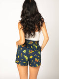 This photo showcases a white female presenting model wearing a white tank top and the Summer Night shorts facing away from the camera. Shorts are a dark blue with yellow and gold colored insects including moths, and beetles. Also includes flowers